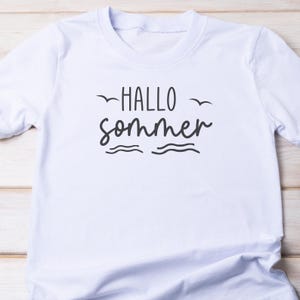 Puede incluir: Camiseta blanca con el texto "Hallo Sommer" en letras negras. El texto est&aacute; estilizado con una l&iacute;nea ondulada debajo y dos p&aacute;jaros volando arriba.