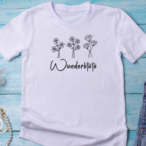 Pu&ograve; includere: T-shirt bianca con un design floreale e la scritta "Wunderblute" in nero. La grafica presenta tre bouquet di fiori stilizzati. La maglietta ha maniche corte e girocollo.