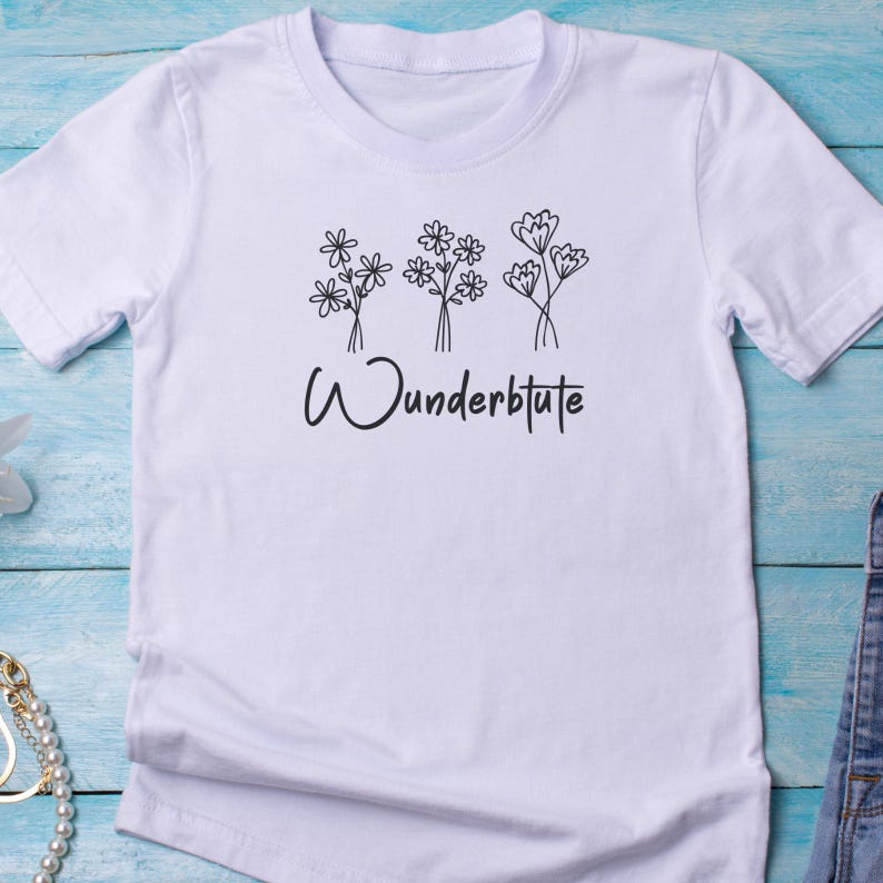 Pu&ograve; includere: T-shirt bianca con un design floreale e la scritta "Wunderblute" in nero. La grafica presenta tre bouquet di fiori stilizzati. La maglietta ha maniche corte e girocollo.