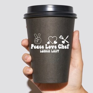 May include: A black to-go coffee cup with a white design that says "Peace Love Chef Lunch Lady" with a peace sign, heart, and kitchen utensils.
