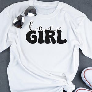 May include: White t-shirt with black text that says "loc girl" in a retro font.