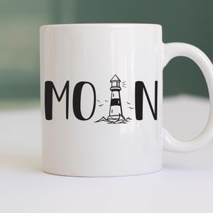 Puede incluir: Taza de cer&aacute;mica blanca con un gr&aacute;fico de faro negro y la palabra "MOAN" impresa en ella.