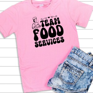 May include: Pink t-shirt with a black graphic that says "Team Food Services" with a cartoon chef holding a plate of food.