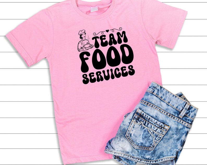 May include: Pink t-shirt with a black graphic that says "Team Food Services" with a cartoon chef holding a plate of food.