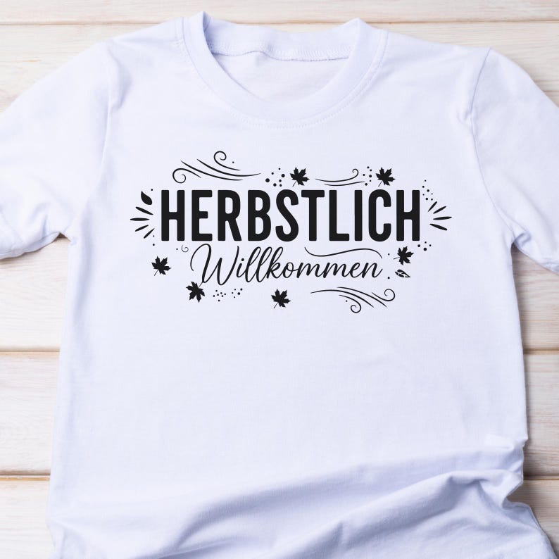 May include: White t-shirt featuring the German phrase "HERBSTLICH Willkommen" in black, with leaf and swirl embellishments. The tee is made of a soft fabric, suitable for everyday wear.
