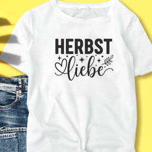 May include: White t-shirt featuring the words "HERBST Liebe" in black. "HERBST" is in a bold, sans-serif font, and "Liebe" is in a cursive font with decorative elements. The shirt is made of a soft material, suitable for casual wear.