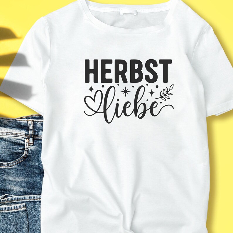 May include: White t-shirt featuring the words "HERBST Liebe" in black. "HERBST" is in a bold, sans-serif font, and "Liebe" is in a cursive font with decorative elements. The shirt is made of a soft material, suitable for casual wear.