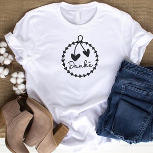 May include: White t-shirt with a black graphic of a wreath with two hearts and the word "Danke" in the center.