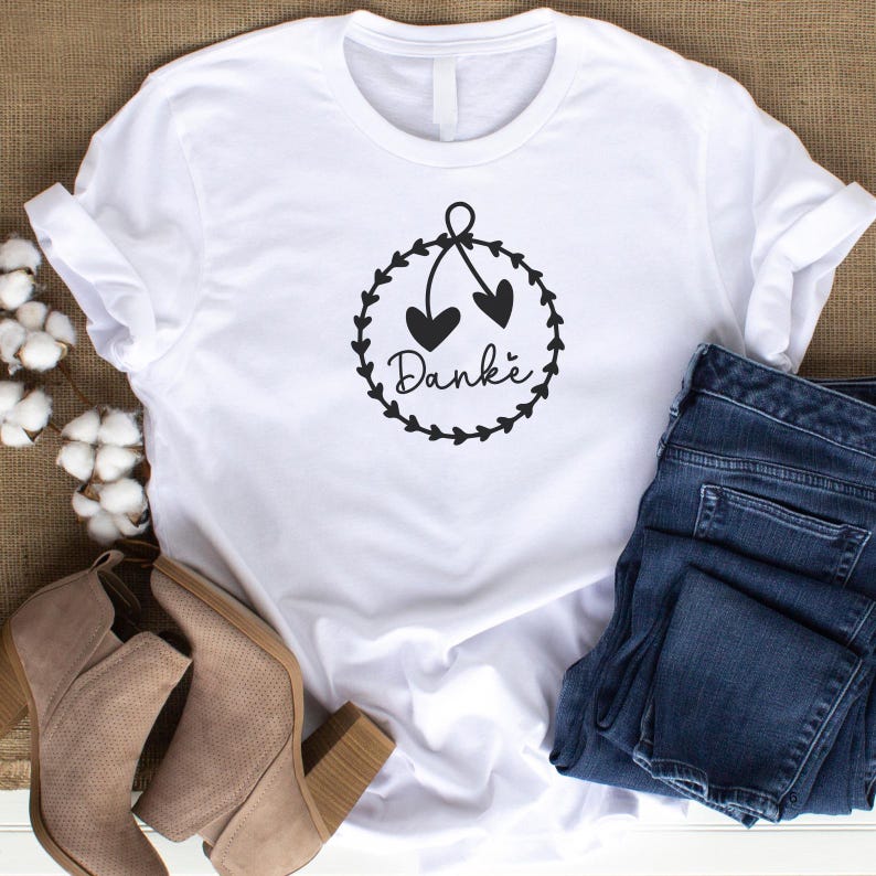 May include: White t-shirt with a black graphic of a wreath with two hearts and the word "Danke" in the center.
