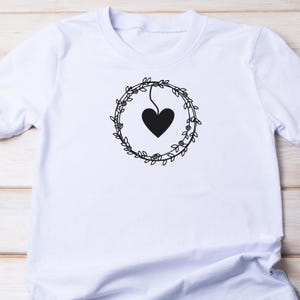 May include: White t-shirt with a black floral wreath design and a heart hanging from the wreath.