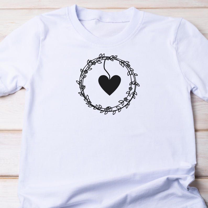 May include: White t-shirt with a black floral wreath design and a heart hanging from the wreath.
