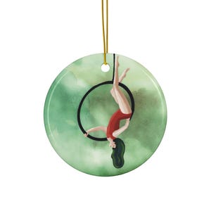 Könnte beinhalten: Ein Keramik-Anhänger mit einer Aquarell-Illustration einer Frau in einem roten Trikot, die an Luftringen an einem grünen Hintergrund performt.