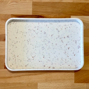 Könnte beinhalten: Ein rechteckiges weißes Tablett mit einem gesprenkelten Design. Das Tablett hat abgerundete Ecken und einen erhöhten Rand. Das gesprenkelte Design weist kleine rosa, braune und weiße Stücke auf. Das Tablett liegt auf einer Holzoberfläche.