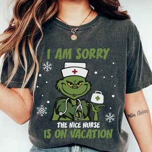 Puede incluir: Camiseta gris oscuro con un personaje similar al Grinch con uniforme de enfermera y el texto "I AM SORRY THE NICE NURSE IS ON VACATION". El diseño incluye copos de nieve y un maletín médico.