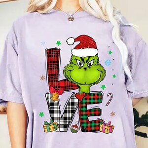 Puede incluir: Una camiseta de color lila claro con un diseño del Grinch con un gorro de Papá Noel. La palabra "LOVE" está escrita con patrones de cuadros y adornos navideños, incluyendo una campana, un corazón y regalos.