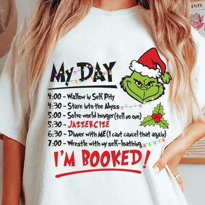 Könnte beinhalten: Weißes T-Shirt mit dem Text "My DAY" und einem humorvollen Zeitplan. Es zeigt einen grünen Grinch-Charakter mit Weihnachtsmütze und den roten Worten "I'M BOOKED!"