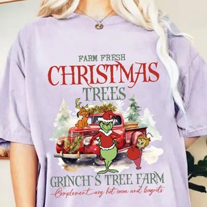 Puede incluir: Una camiseta lila claro con un diseño navideño. El diseño incluye una camioneta roja, el Grinch y el texto "Farm Fresh Christmas Trees" y "Grinch's Tree Farm". El gráfico es en rojo, verde y blanco.