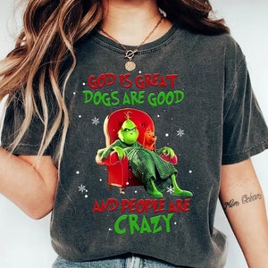 Puede incluir: Camiseta gris oscuro con un diseño temático del Grinch. El Grinch está sentado en una silla roja con un perro, y el texto dice "GOD IS GREAT, DOGS ARE GOOD, AND PEOPLE ARE CRAZY" en rojo y verde.