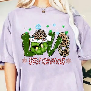 Puede incluir: Camiseta lila claro con un diseño gráfico. La palabra "LOVE" está en verde y estampado de leopardo, con la "O" como un personaje verde de Grinch con un gorro de Papá Noel. "Grinchmas" está escrito en rayas rojas y blancas de bastón de caramelo.