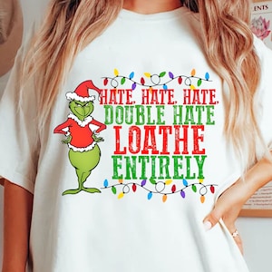 Puede incluir: Camiseta blanca con un personaje de dibujos animados verde con gorro de Papá Noel y atuendo rojo. La camiseta tiene el texto "HATE, HATE, HATE, DOUBLE HATE, LOATHE ENTIRELY" en verde y rojo, con una guirnalda de luces navideñas.