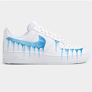 May include: White leather sneaker with a blue swoosh logo and blue icicle design. The shoe features a white sole and laces, with the word "AIR" printed on the side.