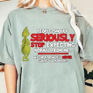 May include: Sage green t-shirt featuring a Grinch cartoon character and the text "PEOPLE SHOULD SERIOUSLY STOP EXPECTING NORMAL FROM ME... we all know it's NEVER going to happen!" in red and white.