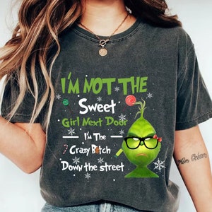 May include: Dark grey t-shirt with the text "I'M NOT THE Sweet Girl Next Door I'M The Crazy Bitch Down the street". The shirt features a green Grinch character wearing glasses and a bow.