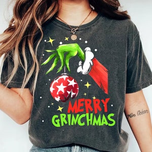 Peut inclure: T-shirt gris foncé avec un motif sur le thème du Grinch. Le motif comprend une main verte tenant une boule rouge et blanche à motifs d'étoiles, avec les mots "MERRY GRINCHMAS" en rouge et vert.