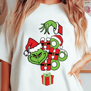 Puede incluir: Camiseta blanca con un personaje verde del Grinch con un gorro de Papá Noel y las palabras "HO HO HO" en verde. Una caja de regalo roja con un lazo verde cuelga debajo. La mano verde del Grinch está en la parte superior.