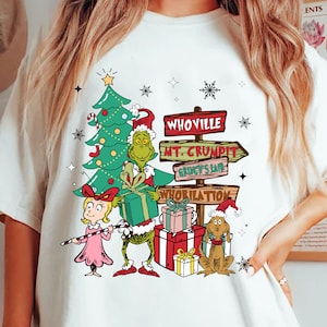 Puede incluir: Camiseta blanca con un diseño navideño. El diseño incluye un árbol de Navidad verde, personajes, cajas de regalo y señales direccionales hacia Villa Quien, Mt. Crumpit y Grinch's Lane. El texto "Whobilation" también es visible.