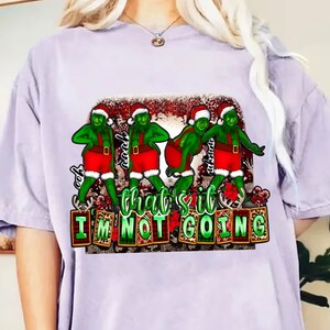 Puede incluir: Una camiseta de color lila claro con un gráfico de cuatro personajes verdes con gorros de Papá Noel y trajes rojos. El texto "that's it I'm not going" se muestra en una fuente festiva, con un tema navideño.