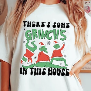 Puede incluir: Camiseta blanca con un gráfico que presenta a tres Grinches verdes con trajes y gorros de Papá Noel rojos. El texto "THERE'S SOME GRINCH'S IN THIS HOUSE" está impreso encima y debajo de los Grinches en negro y verde.
