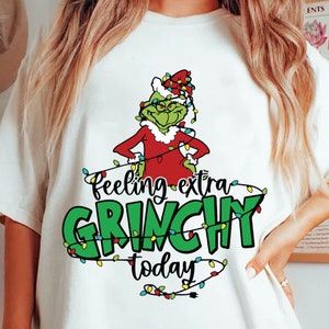 Könnte beinhalten: Weißes T-Shirt mit einem Grinch-Charakter in rotem Weihnachtsmann-Anzug und -Hut, umgeben von Lichterketten. Der Text lautet "Feeling extra Grinchy today" in Grün und Schwarz.