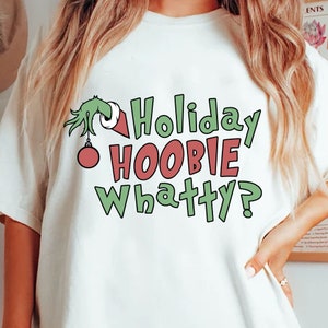 May include: A cream-coloured t-shirt with a green hand holding a red and green bauble. The shirt displays the text "Holiday HOOBIE Whatty?" in green and red lettering, suggesting a festive holiday theme.