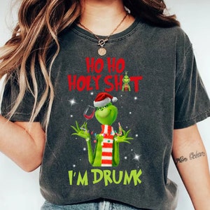 Puede incluir: Camiseta gris oscuro con un personaje verde de Grinch con un gorro de Papá Noel, sosteniendo una copa de vino y un bastón de caramelo. El texto "HO HO HOLY SHIT I'M DRUNK" está impreso en rojo y verde.