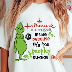 Puede incluir: Camiseta blanca con un personaje verde de Grinch y el texto "Hallmark Christmas Movies inside because it's too peopley outside." El diseño incluye una corona roja, copos de nieve y hojas verdes.