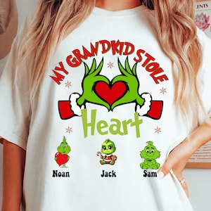 Puede incluir: Camiseta blanca con un diseño temático de Grinch. El texto dice "My Grandkid Stole Heart" con manos de Grinch formando un corazón. Debajo hay tres personajes de Grinch con los nombres "Noan", "Jack" y "Sam".
