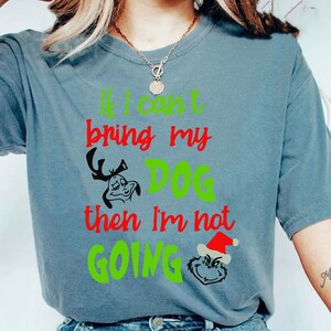 Puede incluir: Camiseta azul grisáceo con el texto "If I can't bring my DOG then I'm not GOING" en verde y rojo. La camiseta presenta ilustraciones de dibujos animados de un personaje parecido al Grinch con cuernos y una cara del Grinch con un gorro de Papá Noel.