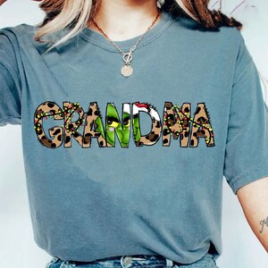 Puede incluir: Camiseta azul grisáceo con la palabra "GRANDMA" en fuente de estampado de leopardo. La letra "A" es reemplazada por una cara verde del Grinch con un gorro de Papá Noel. Las letras están decoradas con luces navideñas.