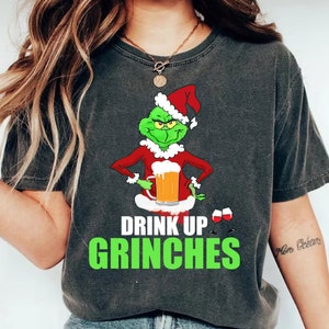 Puede incluir: Camiseta gris oscuro con un personaje de dibujos animados del Grinch con un gorro de Papá Noel y un traje rojo, sosteniendo una jarra de cerveza. El texto "DRINK UP GRINCHES" está impreso en verde y blanco.