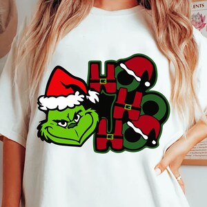 Puede incluir: Camiseta blanca con un personaje verde de Grinch con un gorro de Papá Noel, y las palabras "HO HO HO" en un diseño festivo. Las letras son rojas con contornos verdes y detalles de gorro de Papá Noel, ideal para Navidad.