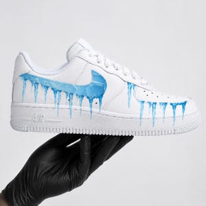 May include: White athletic shoe with a blue swoosh and blue icicle design. The shoe has the word "AIR" on the side. The shoe is held by a hand wearing a black glove.
