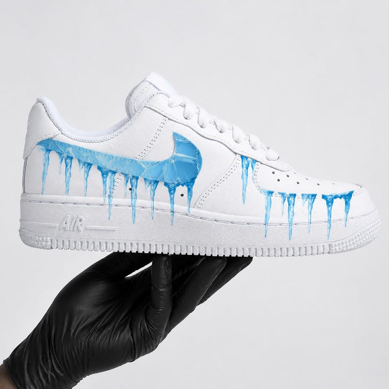 May include: White athletic shoe with a blue swoosh and blue icicle design. The shoe has the word "AIR" on the side. The shoe is held by a hand wearing a black glove.