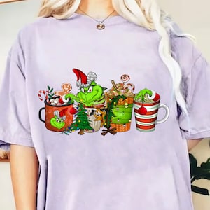 Puede incluir: Camiseta de color lila claro con un diseño navideño que presenta al Grinch y tazas festivas. Las tazas están decoradas con bastones de caramelo, adornos y un hombre de jengibre. El Grinch lleva un gorro de Papá Noel.
