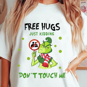 Puede incluir: Camiseta blanca con un gráfico del Grinch sosteniendo un letrero de "sin abrazos". El texto dice "FREE HUGS JUST KIDDING DON'T TOUCH ME". El Grinch lleva una bufanda a rayas rojas y blancas.