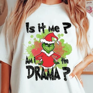Puede incluir: Camiseta blanca con un personaje de Grinch con gorro de Papá Noel y atuendo rojo, con el texto "Is it me? Am I the DRAMA?" en negro. El diseño incluye salpicaduras de pintura verde y roja.