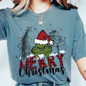 Puede incluir: Camiseta azul verdoso con un gráfico del Grinch con un gorro de Papá Noel y las palabras "Merry Christmas" en un patrón de cuadros rojos y negros. También incluye un árbol de Navidad y copos de nieve.
