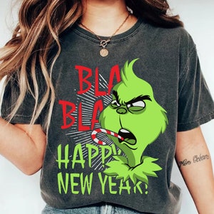 Puede incluir: Camiseta gris oscuro con un personaje verde del Grinch, un bastón de caramelo y el texto "BLA BLA HAPPY NEW YEAR!" en rojo y verde. El Grinch tiene una expresión enfadada. Un diseño festivo para el Año Nuevo.
