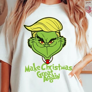 Puede incluir: Una camiseta blanca con un personaje verde parecido al Grinch con pelo amarillo y ojos rojos. El personaje lleva una corbata roja. El texto "Make Christmas Great Again" está impreso debajo del personaje.