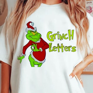 May include: White t-shirt with a cartoon Grinch character wearing a Santa hat and red coat with white trim. The Grinch is green with yellow eyes and is gesturing with his hand. The words "Grinch Letters" are in green, outlined in black.